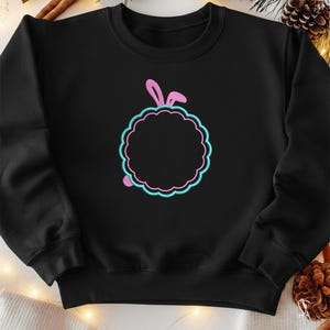 Puede incluir: Sudadera negra con un diseño de círculo festoneado rosa y turquesa y orejas de conejo rosas. El diseño está centrado en la parte delantera de la sudadera. La sudadera es de un material suave.