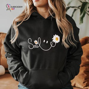May include: Black hoodie with a white "Smile" design. The design features the word "Smile" in cursive, with a daisy flower replacing the "i" and another daisy forming the right side of the smile. The hoodie has a front pocket and a drawstring hood.