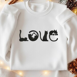 May include: White sweatshirt with the word "LOVE" embroidered in black. The "O" is a circle with two cats and a heart. The other letters are stylized with cat silhouettes. A cozy, casual top for cat lovers.