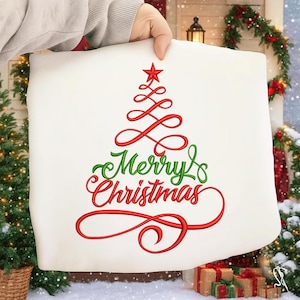 Op de afbeelding: Een wit kussen met een rood geborduurd kerstboomontwerp en de woorden "Merry Christmas" in rode en groene letters. Het kussen wordt door een hand omhoog gehouden.
