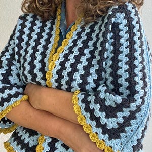 May include: A handmade crochet cardigan in a granny square pattern. The cardigan features a colour block design with navy blue, light blue, and yellow trim. The sleeves are long and the cardigan is open in the front.