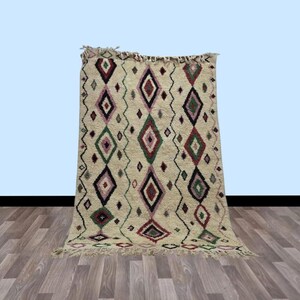 Könnte beinhalten: Cremefarbener Teppich mit einem Rautenmuster in Rot-, Grün- und Lilatönen. Der Teppich hat einen Fransenrand und einen Zickzack-Bordüre. Der Teppich wird auf einem Holzboden präsentiert.