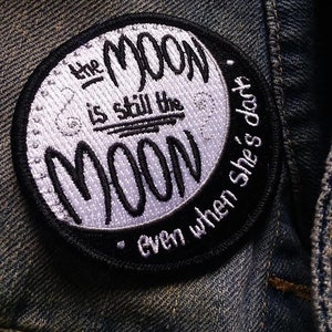May include: A black and white embroidered patch with the text "The Moon is still the Moon, even when she's dark."