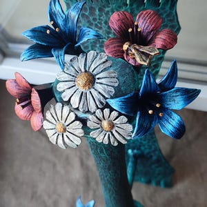 Può includere: Un vaso blu turchese con superficie testurizzata è decorato con una varietà di fiori in pelle. I fiori includono margherite bianche, fiori rossi e rosa e gigli blu. Una piccola ape dorata è posata su uno dei fiori rossi.