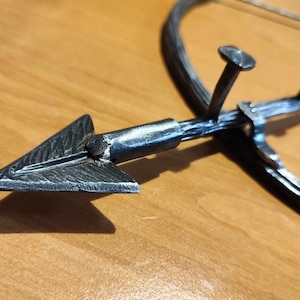 Könnte beinhalten: Ein handgefertigter Metallpfeil mit dreieckiger Pfeilspitze. Der Pfeil ist an einem gebogenen Metallstück mit drei nagelartigen Vorsprüngen befestigt. Das Metall hat ein strukturiertes, rustikales Aussehen.