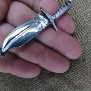 Puede incluir: Un pequeño colgante de daga plateada con una hoja curva y una guarda cruzada. El mango está detallado con líneas horizontales. El colgante tiene un lazo en la parte superior para una cadena. Fondo texturizado marrón.