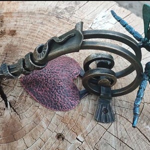 Może przedstawiać: Dekoracyjny metalowy klamka do drzwi z ozdobnym wzorem węża i dwoma akcentami w postaci ważek. Klamka jest wykończona w kolorze brązu, a ważki są niebieskie i zielone.