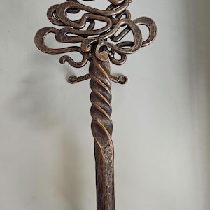Puede incluir: Una llave grande de metal con un diseño intrincado, un eje retorcido y una parte superior decorativa con forma de árbol. La llave está montada en un soporte con dos pequeños bucles.
