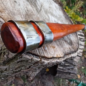 May include: A wooden club with a reddish-brown finish, featuring two silver metal bands around the shaft. The club has a rounded head and a tapered handle. The image is taken outdoors, with the club resting on a tree stump.