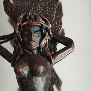 Könnte beinhalten: Eine Bronzeskulptur einer Frau mit langen Haaren, die Arme über dem Kopf erhoben. Die Skulptur ist detailliert mit einer strukturierten Oberfläche und einem Blattdesign.