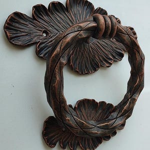 May include: A decorative door knocker made of metal with a twisted rope design and leaf-shaped accents. The knocker is finished in a copper color.