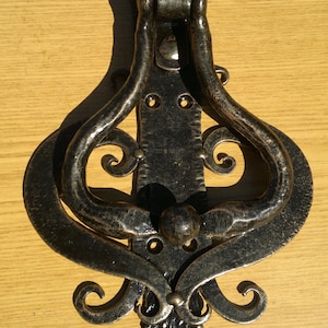 May include: Ornate, dark metal door knocker with a decorative, scroll-like design. The knocker features a central vertical bar with curved arms and a stylized floral base. The finish has a rustic, aged appearance.