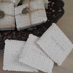 Puede incluir: Una pila de galletas de cerámica blanca con el texto "Petit Beurré" en relieve. Las galletas están atadas con cuerda y tienen un pequeño amuleto de metal adjunto.