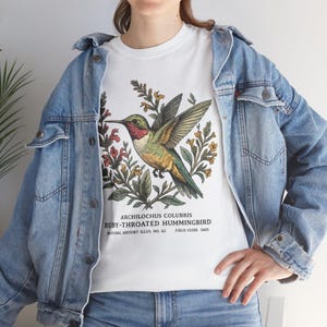 Pode incluir: Camiseta branca com uma ilustração detalhada de um beija-flor-rubi e elementos botânicos. O design inclui o nome científico, "Archilochus Colubris", e texto de um guia de campo. A camiseta é usada com uma jaqueta jeans azul.