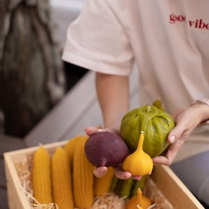 Puede incluir: Una caja de madera llena de velas artesanales con forma de verduras. Las velas son de varios colores, incluyendo verde, amarillo, rojo y morado. Las velas están dispuestas en un lecho de paja. El texto "good vibes" es visible en una camisa blanca.