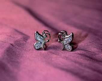 May include: A pair of silver butterfly stud earrings. Each earring features a butterfly design with a body and wings encrusted with small, clear gemstones. The earrings are displayed on a textured, deep pink fabric.