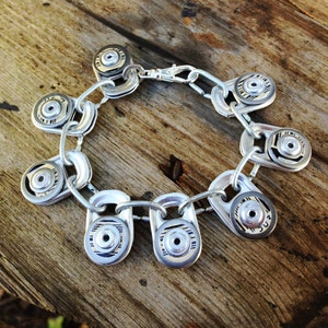 Peut inclure: Un bracelet argenté fait de tirettes de canettes en aluminium recyclées. Le bracelet est composé d'une série de breloques circulaires avec un motif argenté et noir.
