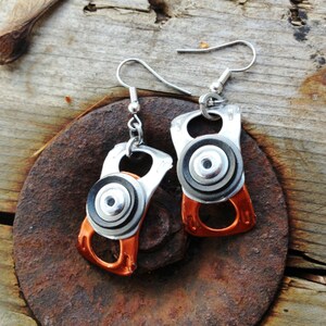 May include: A pair of earrings made from recycled aluminium beverage can tabs. The earrings are silver with black and white accents and have a copper-coloured ring at the top. The earrings are hanging from silver hooks.