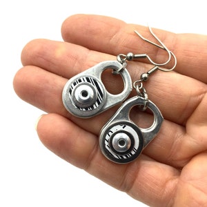 Op de afbeelding: Een paar zilveren oorbellen gemaakt van gerecyclede treklipjes. Elke oorbel heeft een zwart-wit gestreept ontwerp met een zilveren ring.