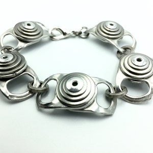 May include: A silver bracelet made from recycled aluminium pull tabs. The bracelet is made of several pull tabs that have been stacked and joined together to create a unique and stylish design.