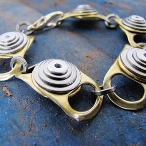 May include: A gold and silver bracelet made from recycled materials. The bracelet features a series of interlocking rings with a circular design.