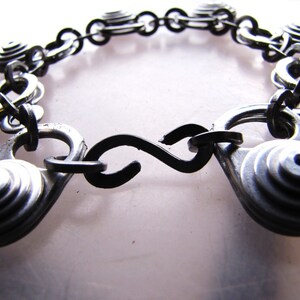 May include: A silver bracelet made from stacked metal rings connected by a black chain. The bracelet has a black metal clasp.