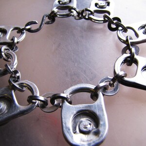 May include: A silver chain bracelet made from connected pull tabs from aluminum cans. The bracelet is made of multiple pull tabs connected together with metal rings.