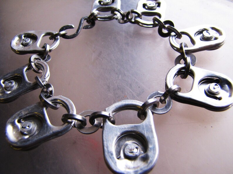 May include: A silver chain bracelet made from connected pull tabs from aluminum cans. The bracelet is made of multiple pull tabs connected together with metal rings.