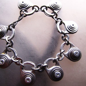 May include: A silver bracelet made from pull tabs from beverage cans. The bracelet is made of a chain with multiple pull tabs attached to it. Each pull tab has a circular design with concentric circles.