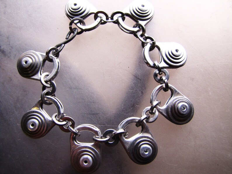 May include: A silver bracelet made from pull tabs from beverage cans. The bracelet is made of a chain with multiple pull tabs attached to it. Each pull tab has a circular design with concentric circles.