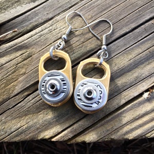 Peut inclure: Une paire de boucles d'oreilles dorées faites à partir de languettes de canettes de soda recyclées. Les boucles d'oreilles ont des crochets argentés et un anneau argenté autour de la languette. Les languettes ont un code-barres noir et blanc.
