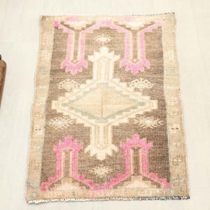 May include: A small, rectangular rug with a geometric pattern. The rug features a central diamond shape and repeating pink and beige motifs on a brown background. The rug's edges are fringed, and it appears to be made of woven material.