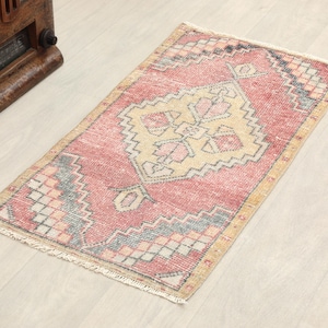 May include: A rectangular, vintage-style rug with a central geometric design. The rug features a faded red background with a beige and pink central pattern. The border includes blue and gray accents, and the rug has a fringed edge.