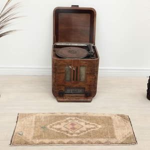 May include: A rectangular rug featuring a tan and beige color palette with a central diamond-shaped geometric pattern. The rug has a short pile texture and a dark brown border, positioned in front of a vintage wooden radio and record player console.