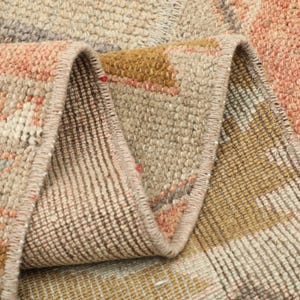 May include: A woven rug featuring a geometric pattern in shades of peach, beige, mustard yellow, and muted green. The rug is folded over to reveal the textured underside, showing the dense pile construction and the finished stitched edges.