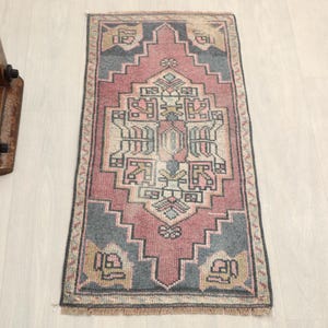 May include: A rectangular, vintage-style rug with a central geometric design in shades of pink, beige, and blue-gray. The rug has a fringed edge and is likely made of woven material. The rug's dimensions are approximately 24 x 48 inches.