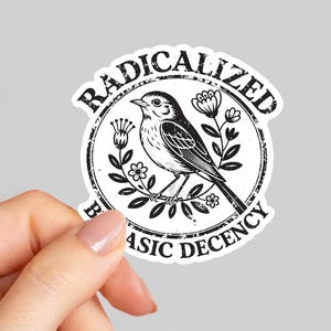 May include: A white sticker with a black and white illustration of a bird perched on a branch, surrounded by flowers. The sticker has the words "RADICALIZED" and "BASIC DECENCY" in a circular design.