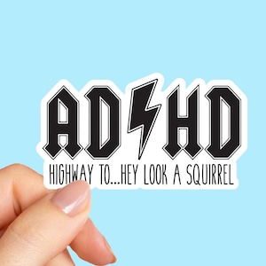 May include: A white sticker with black text that reads "ADHD" in a bold, stylized font, with a lightning bolt in the center. Below, the text continues "HIGHWAY TO...HEY LOOK A SQUIRREL". The sticker is held against a light blue background.
