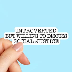 May include: A white sticker with black text that reads "INTROVERTED BUT WILLING TO DISCUSS SOCIAL JUSTICE" is held against a light blue background. The sticker has a black border and is held by a hand.