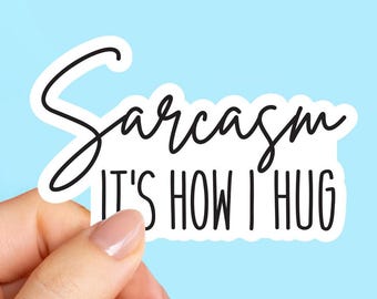 May include: A white sticker with the text "Sarcasm It's How I Hug" in black script and block letters. The sticker is held by a hand against a light blue background.