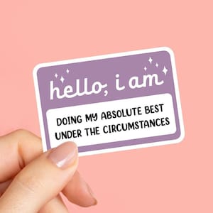 May include: A rectangular sticker with a purple background and white border. The sticker reads "hello, i am" in white script, with the text "DOING MY ABSOLUTE BEST UNDER THE CIRCUMSTANCES" below. The sticker is held by a hand against a pink background.