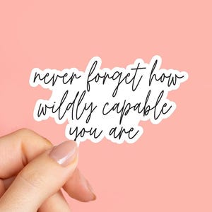 May include: A white sticker with black cursive text that reads "never forget how wildly capable you are." The sticker is held against a pink background.