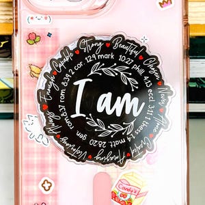May include: A pink phone case with a clear border, decorated with stickers including a bow, bunny, and a candy graphic. The case features the words "I AM" in a circular design with inspirational words.