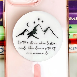 May include: A pink phone case with a white pop socket featuring a mountain and stars design, with the text "to the stars who listen and the dreams that are answered." The phone case is on top of a stack of books.