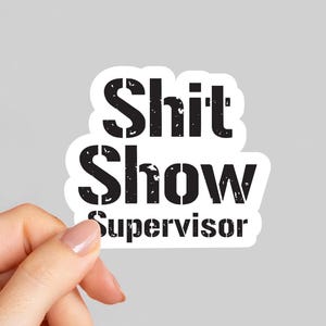 May include: A white sticker with the words "Shit Show Supervisor" in distressed black font. The sticker is held by a hand against a light gray background.