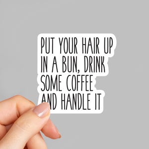 May include: A white sticker with black text that reads: "PUT YOUR HAIR UP IN A BUN, DRINK SOME COFFEE AND HANDLE IT." The sticker is held by a hand against a gray background.