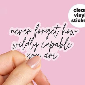 May include: A clear vinyl sticker with the text "never forget how wildly capable you are" in black script. The sticker is held against a pink background. A white circle with the words "clear vinyl sticker" is in the upper right corner.