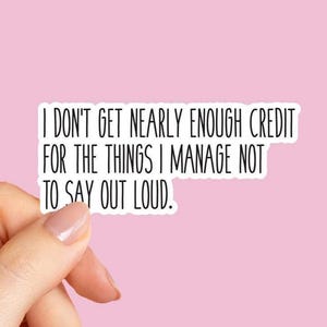 May include: A white sticker with black text that reads, "I DON'T GET NEARLY ENOUGH CREDIT FOR THE THINGS I MANAGE NOT TO SAY OUT LOUD." The sticker is held by a hand against a pink background.