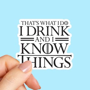 May include: White sticker with black text that reads "THAT'S WHAT I DO I DRINK AND I KNOW THINGS" on a light blue background. The sticker is held by a hand.