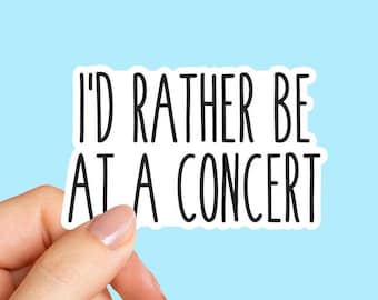 May include: A white sticker with black text that reads "I'D RATHER BE AT A CONCERT" is held by a hand against a light blue background. The sticker has a black border.